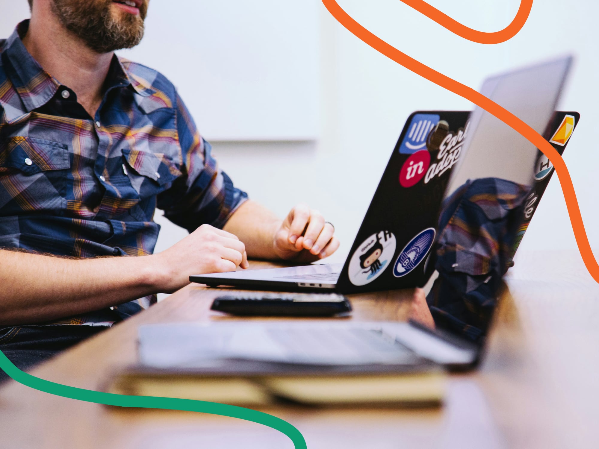 Ein Mann mit Bart und kariertem Hemd sitzt an einem Holztisch und arbeitet an einem Laptop mit mehreren Aufklebern. Im Vordergrund sind ein weiteres Notebook, ein Notizbuch und eine Fernbedienung zu sehen.