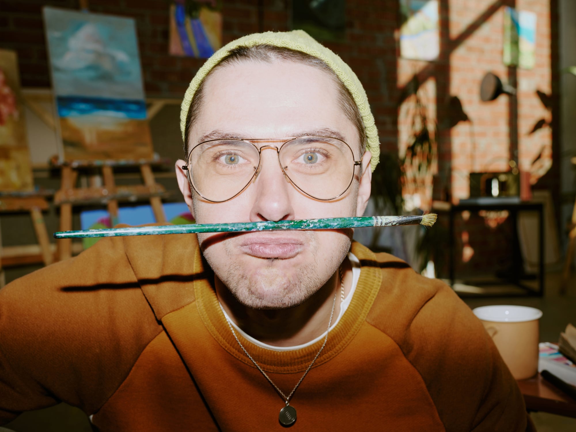 Ein Mann mit heller Haut, Brille und grünem Beanie hält einen Pinsel horizontal unter seiner Nase und macht eine spielerische Grimasse. Er trägt ein braunes Sweatshirt und eine silberne Kette. Im Hintergrund steht eine Staffelei mit mehreren Leinwänden, die bunte Gemälde zeigen. Der Raum ist ein helles Atelier mit Backsteinwänden und großen Fenstern, durch die Sonnenlicht einfällt.